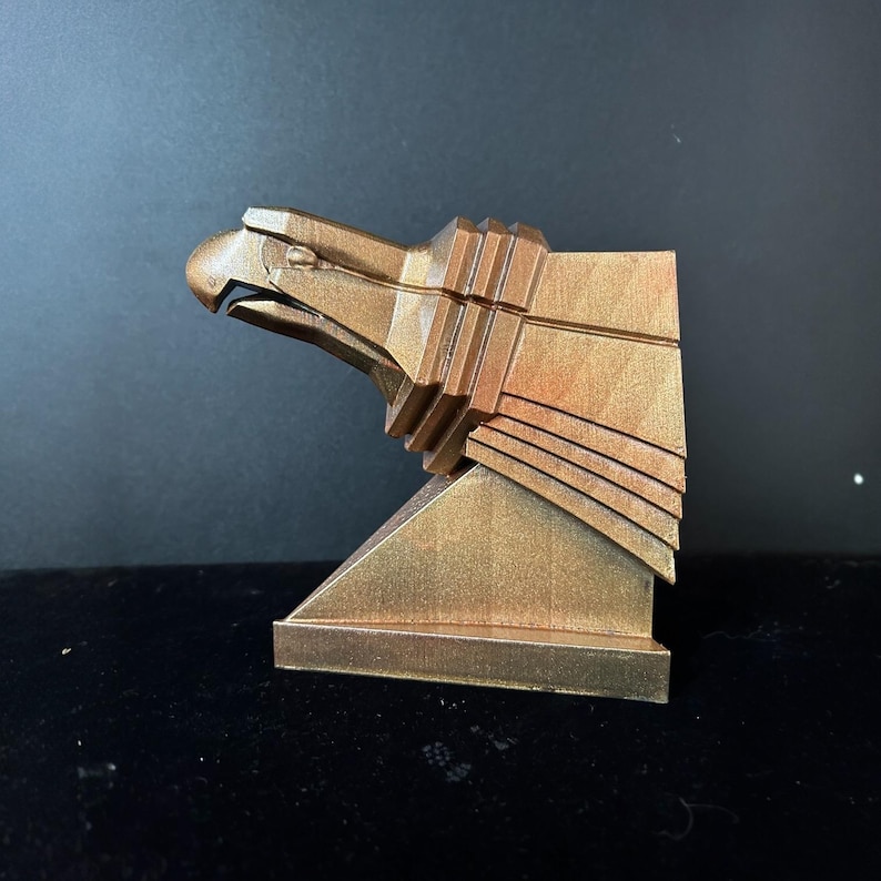 May include: A gold-colored eagle sculpture with a geometric design. The eagle's head is turned to the side, and its wings are spread out. The sculpture is mounted on a square base.