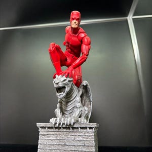 Può includere: Una action figure rossa di un supereroe in posa dinamica, appollaiata su una statua di gargoyle grigia e dettagliata. La figura indossa un completo rosso con una maschera. Il gargoyle è su una base di pietra.