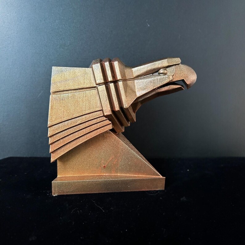 May include: A bronze-colored eagle sculpture with a geometric design. The eagle's head is turned to the right, and its wings are spread out. The sculpture is mounted on a rectangular base.