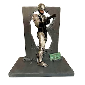 Può includere: Una action figure di un robot argentato in piedi in un muro rotto con una pistola in mano. La figura tiene una pistola e sta in piedi in un muro rotto. La figura indossa un casco e ha un corpo argentato. La figura è in piedi su una base grigia con un cartello verde che dice "Benvenuti a Detroit".