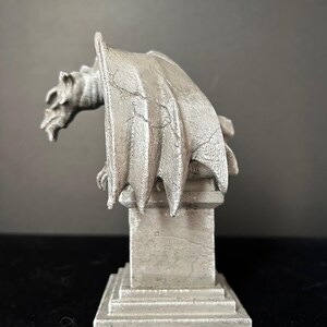 K&ouml;nnte beinhalten: Eine silberfarbene Gargoyle-Statue mit detaillierten Fl&uuml;geln und einem strengen Ausdruck. Die Statue thront auf einem quadratischen Sockel und zeigt komplizierte Texturen und eine gotische &Auml;sthetik. Die Skulptur ist ein dekorativer Artikel.