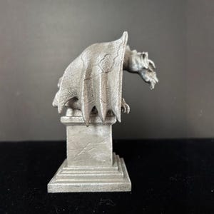 K&ouml;nnte beinhalten: Eine detaillierte, silberfarbene Gargoyle-Statue mit ausgebreiteten Fl&uuml;geln, die auf einem quadratischen Sockel sitzt. Die Statue hat eine strukturierte Oberfl&auml;che und ist vor einem dunklen Hintergrund platziert. Die Gargoyle ist ein dekoratives Objekt.
