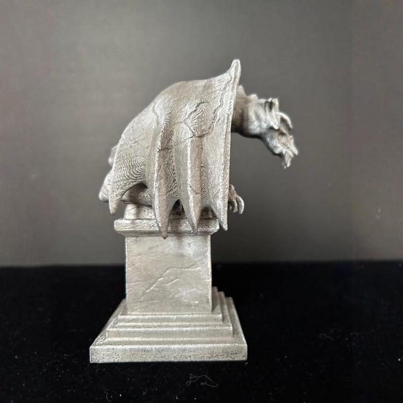 K&ouml;nnte beinhalten: Eine detaillierte, silberfarbene Gargoyle-Statue mit ausgebreiteten Fl&uuml;geln, die auf einem quadratischen Sockel sitzt. Die Statue hat eine strukturierte Oberfl&auml;che und ist vor einem dunklen Hintergrund platziert. Die Gargoyle ist ein dekoratives Objekt.