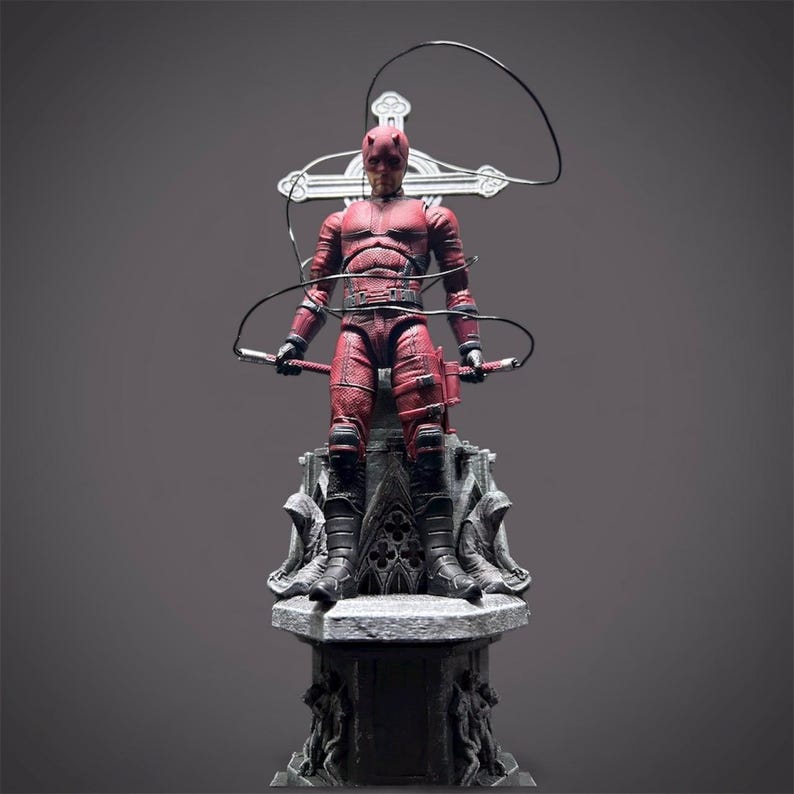 May include: A red action figure of a superhero, posed on a detailed gray pedestal. The figure is wearing a textured red suit, black boots, and has horns. It holds two batons and a wire. A cross-shaped structure is behind the figure.