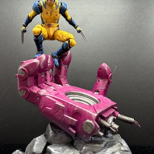Può includere: Una action figure di Wolverine in costume giallo e blu con artigli estesi, appollaiata su una grande mano robotica rosa. La mano poggia su una base rocciosa grigia. La figura è un oggetto da collezione.