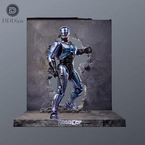 May include: A Robocop action figure with a metallic blue and purple finish, standing in a dynamic pose against a cracked concrete wall backdrop. The base features the text ROBOCOP in silver lettering, and the figure wears a full-body suit.