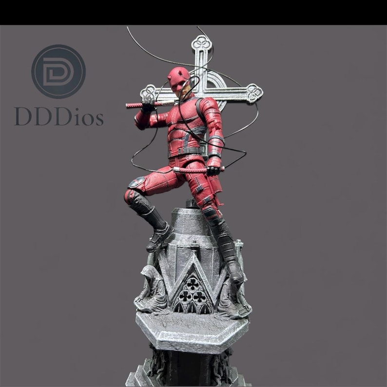 May include: A red action figure of a superhero in a dynamic pose, holding a weapon, atop a detailed stone structure. The figure is clad in a textured red suit with black boots. A cross-shaped emblem is behind the figure.
