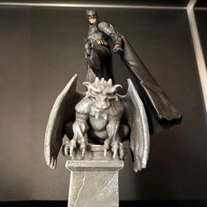 Può includere: Una action figure di Batman è in piedi sopra una statua di gargoyle grigia. La figura di Batman indossa un costume nero e grigio con un lungo mantello nero. Il gargoyle ha grandi ali ed è appollaiato su un piedistallo di pietra. Lo sfondo è scuro.