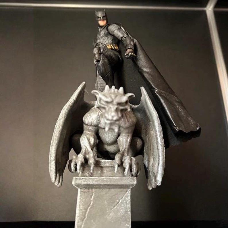 K&ouml;nnte beinhalten: Eine Batman-Actionfigur steht auf einer grauen Gargoyle-Statue. Die Batman-Figur tr&auml;gt einen schwarz-grauen Anzug mit einem langen schwarzen Umhang. Der Gargoyle hat gro&szlig;e Fl&uuml;gel und sitzt auf einem Steinsockel. Der Hintergrund ist dunkel.