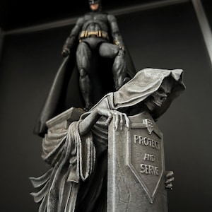 Può includere: Una statua dettagliata con Batman in piedi sopra una figura simile alla Morte. La Morte tiene una lapide con le parole "PROTECT AND SERVE". Le figure sono principalmente grigie con accenti neri, creando un'estetica drammatica.