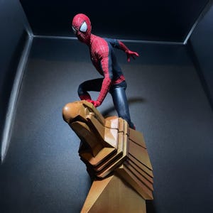 May include: A red and black action figure of Spider-Man standing on a gold eagle statue. The figure is posed in a dynamic action pose with one leg raised and the other foot on the statue.