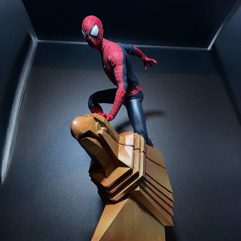 May include: A red and black action figure of Spider-Man standing on a gold eagle statue. The figure is posed in a dynamic action pose with one leg raised and the other foot on the statue.