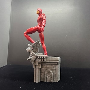 Pode incluir: Uma figura de a&ccedil;&atilde;o vermelha do Daredevil em p&eacute; em um diorama de telhado cinza. O diorama apresenta uma g&aacute;rgula com asas.