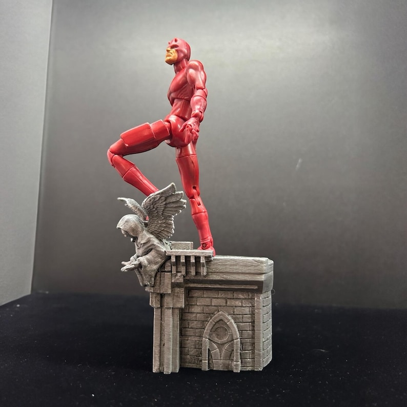 Pode incluir: Uma figura de a&ccedil;&atilde;o vermelha do Daredevil em p&eacute; em um diorama de telhado cinza. O diorama apresenta uma g&aacute;rgula com asas.