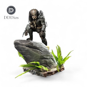 May include: A detailed Predator action figure in a crouching pose, atop a gray rock formation with green foliage. The figure is clad in dark armor and holds a weapon. The logo "DDDios" is in the upper left corner.