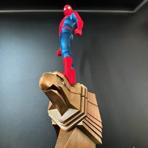 May include: A red and blue superhero figurine standing on a gold eagle statue. The superhero is wearing a red and blue suit with a spider logo on the chest. The eagle statue is detailed and has a sharp beak.