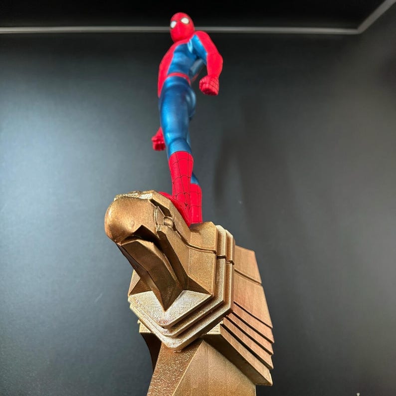 May include: A red and blue superhero figurine standing on a gold eagle statue. The superhero is wearing a red and blue suit with a spider logo on the chest. The eagle statue is detailed and has a sharp beak.