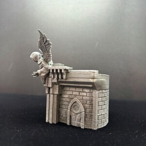May include: A 3D printed miniature of a stone building with a gargoyle on top. The gargoyle is a hooded figure with wings and is facing to the left. The building has a small arched doorway and a brick facade.