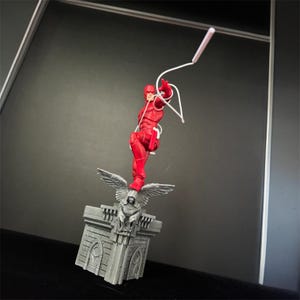 Pode incluir: Uma figura de a&ccedil;&atilde;o vermelha do Daredevil, um super-her&oacute;i, est&aacute; no ar, segurando um bast&atilde;o branco. Ele est&aacute; em p&eacute; sobre uma base esculpida cinza com uma figura de g&aacute;rgola.