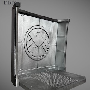 May include: A silver-colored display stand with a circular emblem featuring a stylized bird design. The stand has a textured platform and a rectangular back panel with riveted details. The top left corner has the letters "DDD".