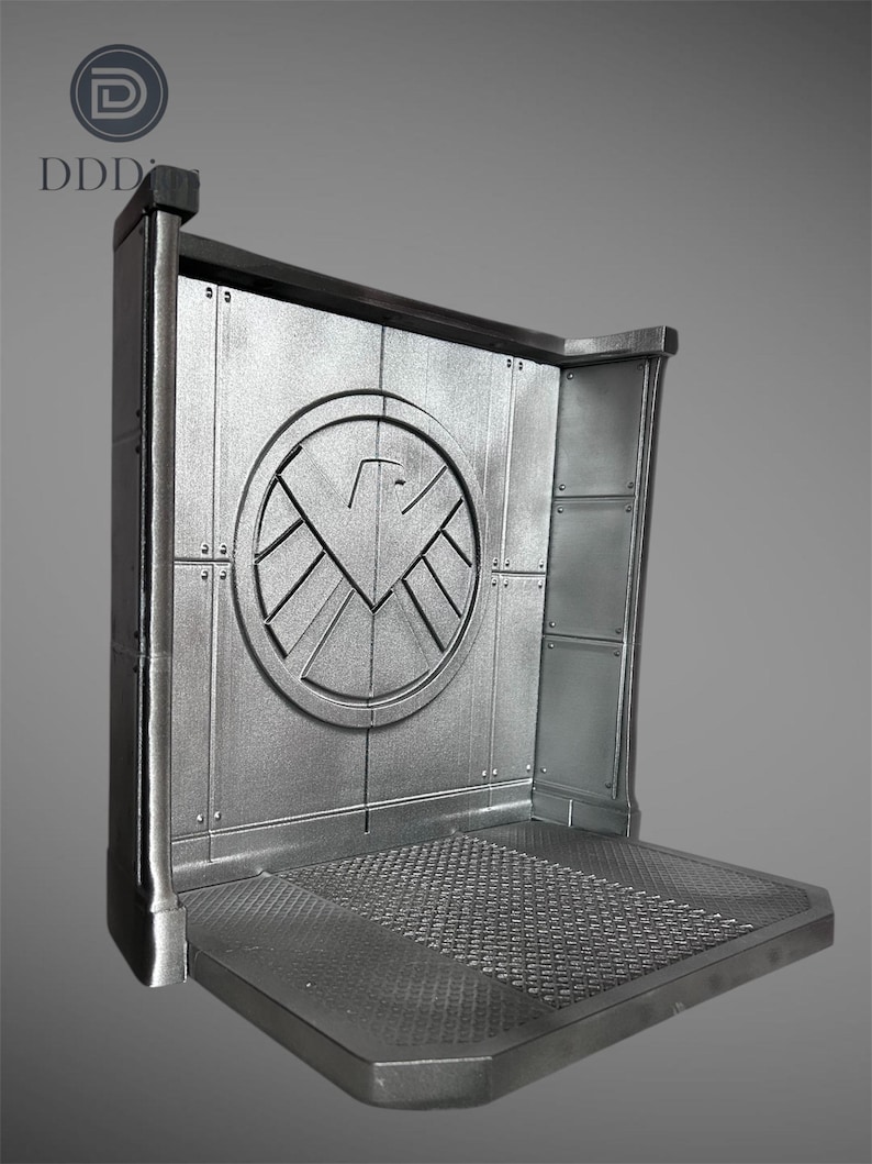 May include: A silver-colored display stand with a circular emblem featuring a stylized bird design. The stand has a textured platform and a rectangular back panel with riveted details. The top left corner has the letters "DDD".