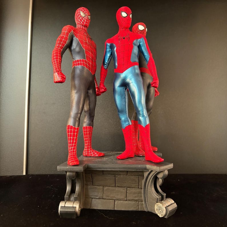 May include: Three action figures of Spider-Man in different poses. The figures are standing on a stone base. The figures are all wearing red and blue suits with black webbing. The figure on the left is wearing a red and black suit with a black mask. The figure in the middle is wearing a blue and red suit with a red mask. The figure on the right is wearing a red and black suit with a black mask.
