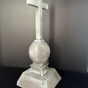 May include: A gray stone cross with a round base and a square base. The cross is on a black background.