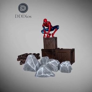 Può includere: Un action figure di Spider-Man in posa accovacciata sopra una pila di tre casse di legno marroni, accompagnata da quattro rocce grigie testurizzate e un piccolo pallet marrone. Il logo DDDios è visibile nell'angolo in alto a sinistra.