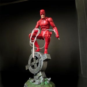Può includere: Una action figure rossa di Daredevil, un supereroe noto per il suo costume rosso e le sue mazze, è appollaiata su una croce di pietra. La figura è in posizione di combattimento, con una mazza tenuta nella mano destra.