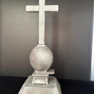 May include: A gray stone sculpture of a cross on top of a sphere, which is on top of a square base.