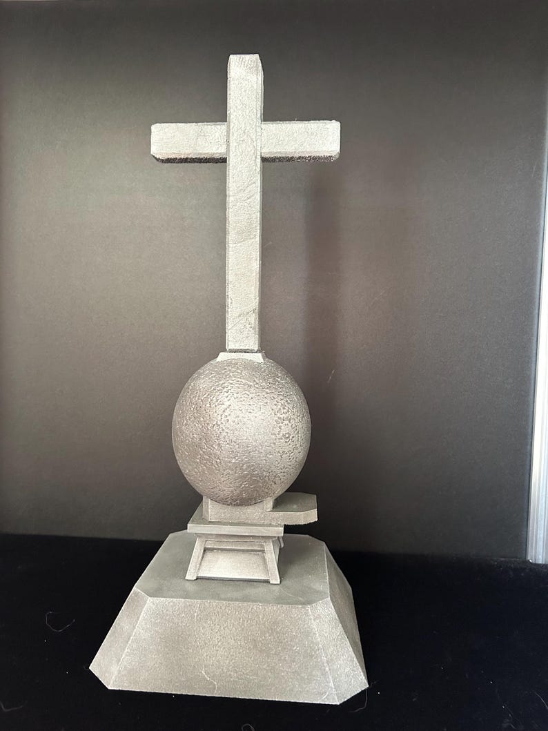 May include: A gray stone sculpture of a cross on top of a sphere, which is on top of a square base.