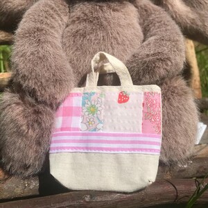 May include: A small, patchwork tote bag with a cream-colored base and a variety of pink and floral patterned panels. The bag features a strawberry applique and a striped section. The bag has a cream-colored handle.