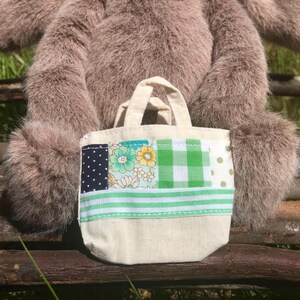 May include: A small, beige tote bag with a patchwork design. The patchwork includes navy blue polka dots, floral patterns, green gingham, and green stripes. The bag has two small handles and is sitting in front of a stuffed animal.