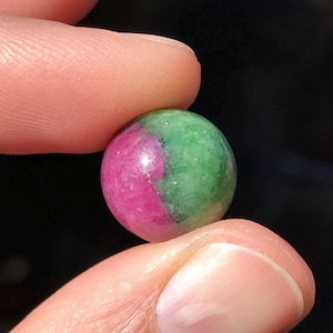 May include: A small, round gemstone held between two fingers. The stone is divided into two colors: a vibrant pink-red and a deep green. The polished surface reflects light, highlighting the contrasting hues. The gemstone's spherical shape is clearly visible.