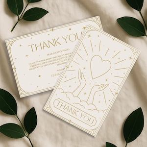 May include: Two ivory-colored thank you cards with gold foil accents. The cards feature the words "THANK YOU" in elegant script, along with a heart and hands design. The cards are decorated with stars and geometric borders. The cards are surrounded by green leaves.
