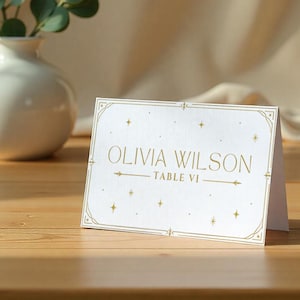 Puede incluir: Una tarjeta de mesa blanca con texto y detalles dorados. La tarjeta dice "OLIVIA WILSON" y "TABLE VI" con flechas decorativas y estrellas. La tarjeta está doblada y sobre una superficie de madera.