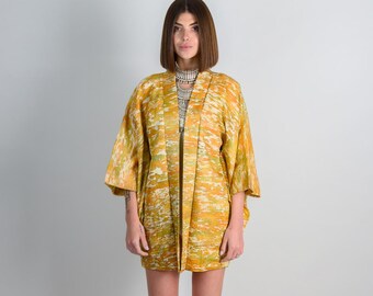 May include: A yellow and white patterned kimono with a long sleeve. The kimono is open in the front and has a silver necklace with a pendant.