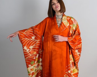 May include: A woman wearing a long, orange kimono with floral patterns. The kimono has a high collar and long sleeves. The woman is wearing a silver necklace and has her hair pulled back.