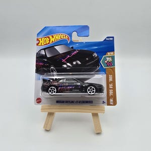 Może przedstawiać: Czarny odlew ciśnieniowy samochodu Nissan Skyline GT-R (BCNR33) Hot Wheels na małym drewnianym sztaludze. Samochód ma białe koła oraz różową i fioletową grafikę. Opakowanie zawiera logo Hot Wheels i tekst "HW: '70s vs. '90s".