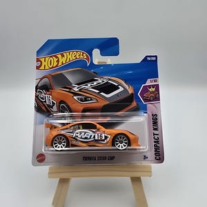 Może przedstawiać: Pomarańczowy odlewany samochód Hot Wheels Toyota GR86 Cup na niebiesko-białej karcie. Samochód ma czarno-białą grafikę z numerem 16. Karta zawiera napisy "Compact Kings" i "79/250".
