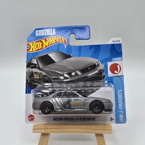 Op de afbeelding: Een zilverkleurige Nissan Skyline GT-R (BCNR33) Hot Wheels gegoten auto, verpakt op een blauw-witte kaart. De kaart toont het Godzilla-logo en de auto-afbeelding. De auto wordt gepresenteerd op een kleine houten ezel.