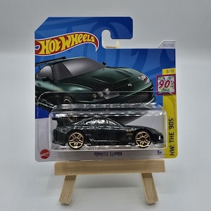 Könnte beinhalten: Ein grünes Hot Wheels-Spielzeugauto aus Druckguss, ein Toyota Supra, mit goldenen Rädern. Das Auto befindet sich auf einer blauen und weißen Karte mit den Worten "Hot Wheels" und "HW: THE '90S" darauf gedruckt. Die Karte hat auch die Nummer "232/250" und "8/10" darauf gedruckt.