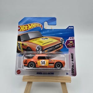 Może przedstawiać: Model samochodu Hot Wheels Honda S800 Racing, pomarańczowy z żółtymi akcentami, z numerem 58. Opakowanie zawiera logo Hot Wheels, tekst "Compact Kings" i numer 212/250.
