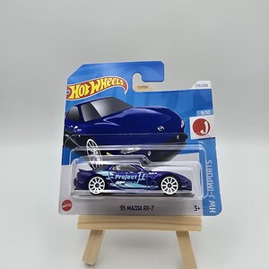 Może przedstawiać: Niebieski samochodzik Hot Wheels '95 Mazda RX-7, prezentowany na małym drewnianym sztaludze. Samochód ma białe koła i grafikę Project ft. Opakowanie zawiera logo Hot Wheels i HW J-Imports.