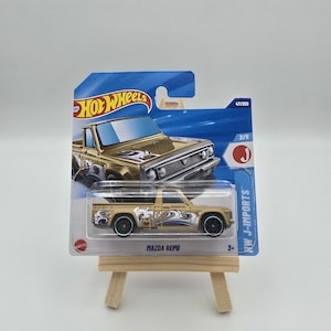 Op de afbeelding: Een goudkleurige Mazda REPU speelgoedtruck met zwarte wielen en blauwe accenten, gepresenteerd op een kleine houten ezel. De verpakking toont het Hot Wheels-logo en de tekst "KW J-IMPORTS".