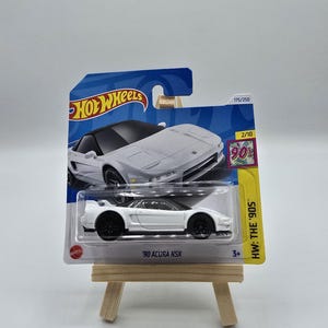 Może przedstawiać: Biały model samochodu '90 Acura NSX Hot Wheels na kartonowym podkładzie. Samochód ma czarne koła i czarny dach. Opakowanie zawiera tekst "HW: THE '90S" i "175/250".