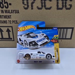 Może przedstawiać: Biały model samochodu Mazda 787B z złotymi kołami, czarnymi i niebieskimi akcentami oraz numerem 18. Samochód jest na niebiesko-żółtej karcie Hot Wheels. Opakowanie zawiera napis "MAZDA 787B".