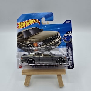 Könnte beinhalten: Ein graues Spielzeugauto '89 Mercedes-Benz 560 SEC AMG aus Druckguss auf einer blau-weißen Hot Wheels-Karte. Das Auto wird auf einer kleinen Holzstaffelei präsentiert. Die Karte trägt das Hot Wheels-Logo und den Text "WILD WIDEBODY".