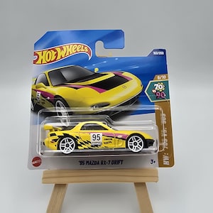Op de afbeelding: Een gele '95 Mazda RX-7 Drift Hot Wheels speelgoedauto op een blauw-witte kaart. De auto heeft zwarte en roze accenten, het nummer 95 en witte wielen. De kaart leest "Hot Wheels" en "9/10".