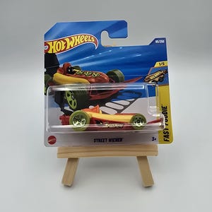 Może przedstawiać: Zabawkowe auto Hot Wheels w przezroczystym plastikowym opakowaniu. Samochód jest czerwono-żółty, z zielonymi kołami, a tekst "STREET WIENER" jest widoczny. Opakowanie ma logo Hot Wheels i numer 85/250.